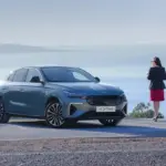 Der neue Togg T10F Fastback steht auf einer Straße mit Blick aufs Meer, daneben eine Frau, Bäume im Hintergrund.