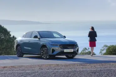 Der neue Togg T10F Fastback steht auf einer Straße mit Blick aufs Meer, daneben eine Frau, Bäume im Hintergrund.