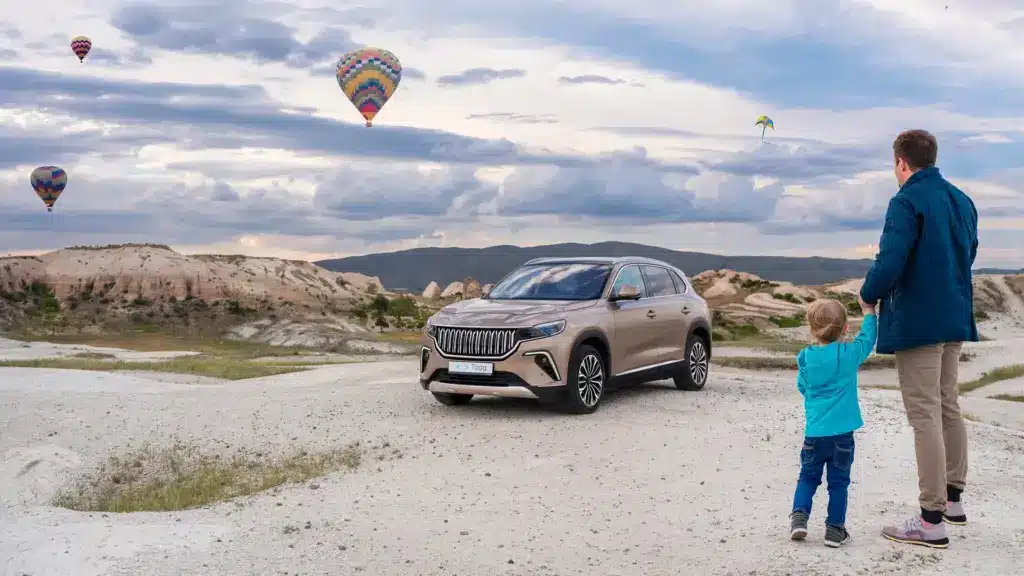 Togg T10X SUV in sandiger Landschaft mit Heißluftballons und zwei Personen im Vordergrund.