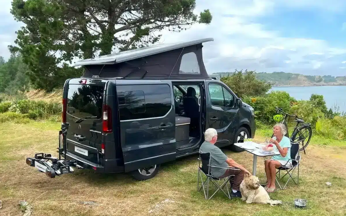 Flex PLUS Campervan mit geöffnetem Aufstelldach, sitzendem Paar und Hund an einem Tisch in der Natur