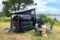 Flex PLUS Campervan mit geöffnetem Aufstelldach, sitzendem Paar und Hund an einem Tisch in der Natur