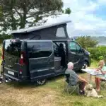 Flex PLUS Campervan mit geöffnetem Aufstelldach, sitzendem Paar und Hund an einem Tisch in der Natur