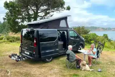 Flex PLUS Campervan mit geöffnetem Aufstelldach, sitzendem Paar und Hund an einem Tisch in der Natur