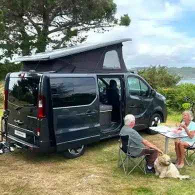 Flex PLUS Campervan mit geöffnetem Aufstelldach, sitzendem Paar und Hund an einem Tisch in der Natur
