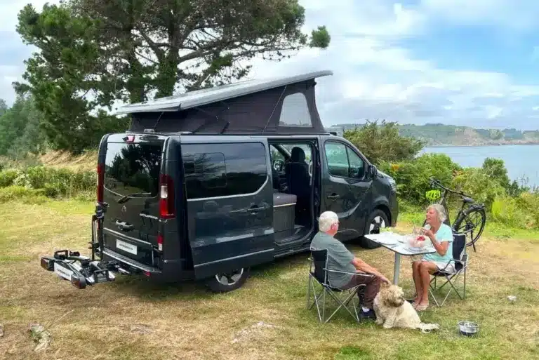 Flex PLUS Campervan mit geöffnetem Aufstelldach, sitzendem Paar und Hund an einem Tisch in der Natur