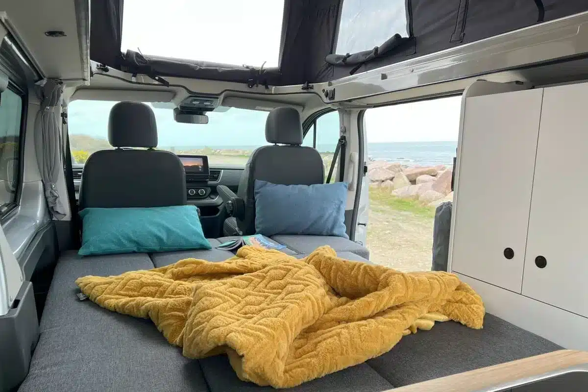 Innenraum des Flex PLUS Campervans mit Schlaffläche, Kissen und Ausblick durch das geöffnete Heck zum Wasser.