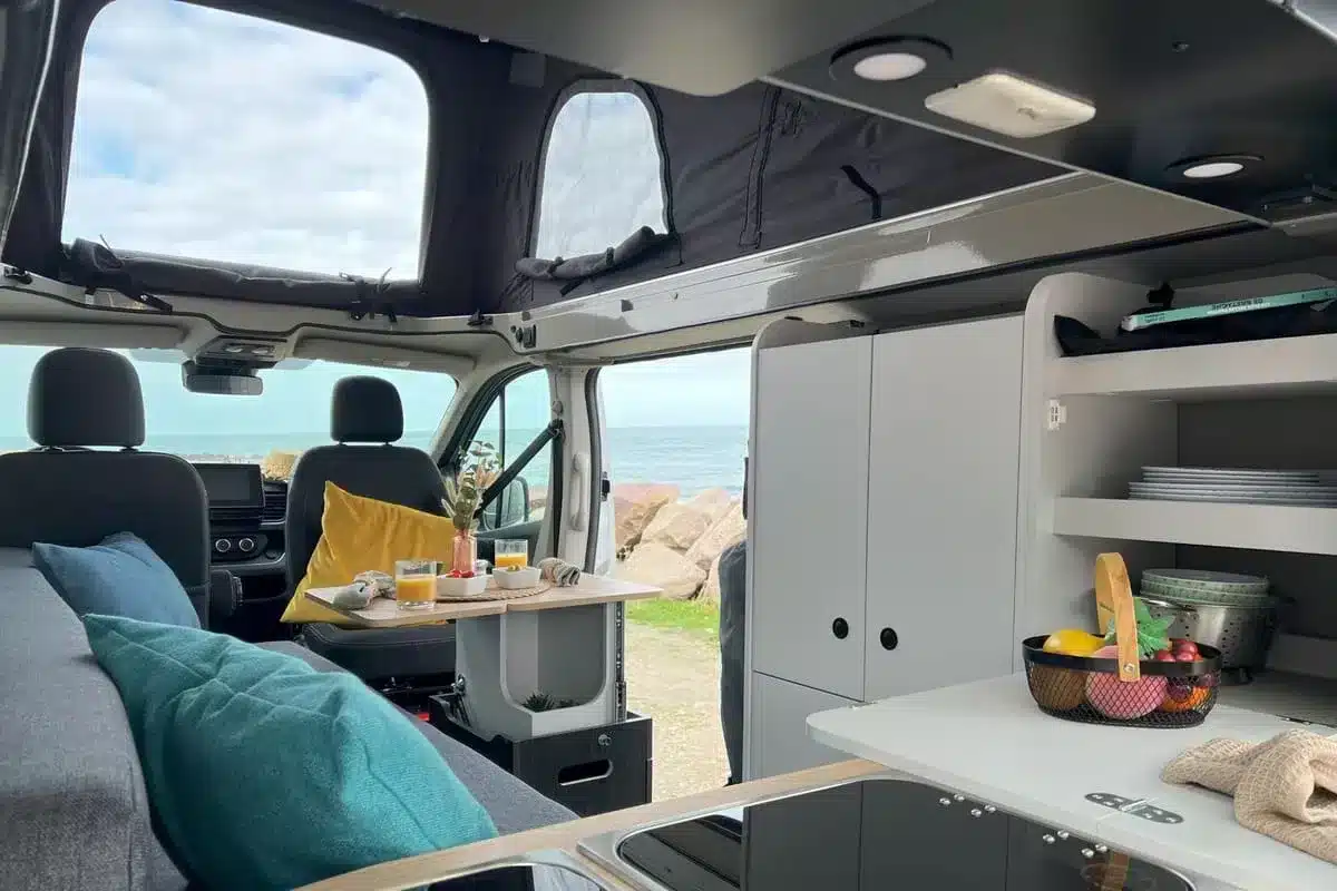 Blick in den Innenraum des Flex PLUS Campervans mit Küche, Tisch und Meer im Hintergrund