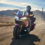 Honda GL1800 Gold Wing Tour 2025 fährt auf einer Landstraße, mit Fahrer und Sozius im Sonnenlicht, Berge im Hintergrund.