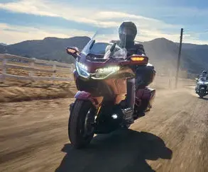 Honda GL1800 Gold Wing Tour 2025 fährt auf einer Landstraße, mit Fahrer und Sozius im Sonnenlicht, Berge im Hintergrund.