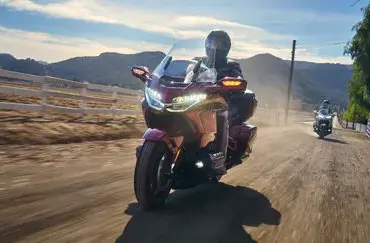 Honda GL1800 Gold Wing Tour 2025 fährt auf einer Landstraße, mit Fahrer und Sozius im Sonnenlicht, Berge im Hintergrund.