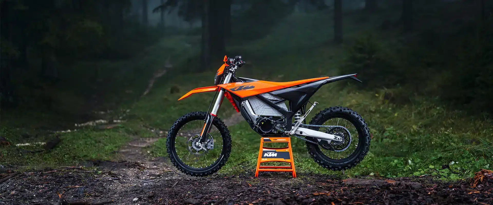 Die 2027 KTM FREERIDE E auf einem Waldweg, seitlich fotografiert, mit markanter oranger Verkleidung und Ständer.