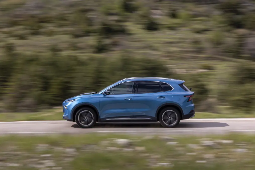Blauer MG SUV fährt auf einer Landstraße durch bewaldete Umgebung, Seitenansicht in Fahrt.