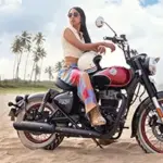 Eine Royal Enfield Goan Classic 350 in Rave Red, gefahren von einer Frau, am Sandstrand mit Palmen und blauem Himmel.