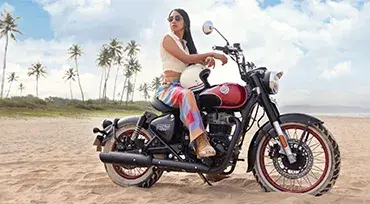 Eine Royal Enfield Goan Classic 350 in Rave Red, gefahren von einer Frau, am Sandstrand mit Palmen und blauem Himmel.