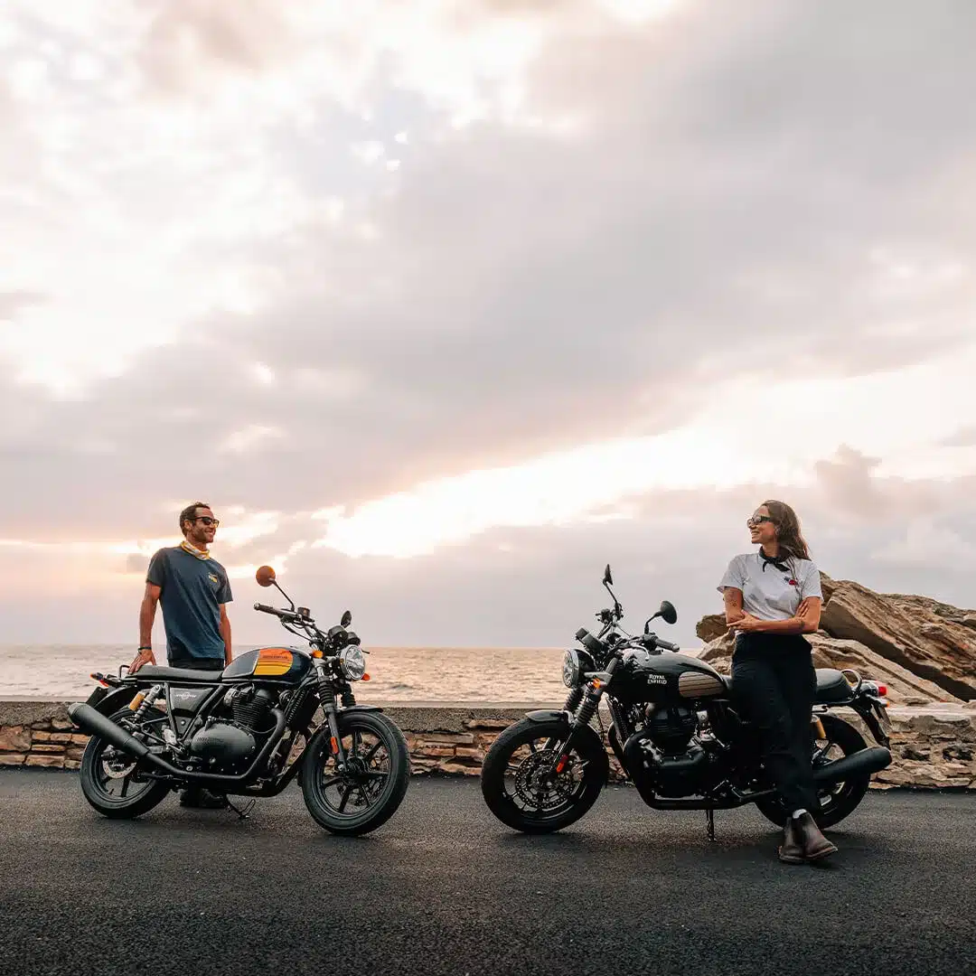 Zwei Royal Enfield Interceptor 650 Motorräder mit zwei Personen am Meer vor Sonnenuntergang.