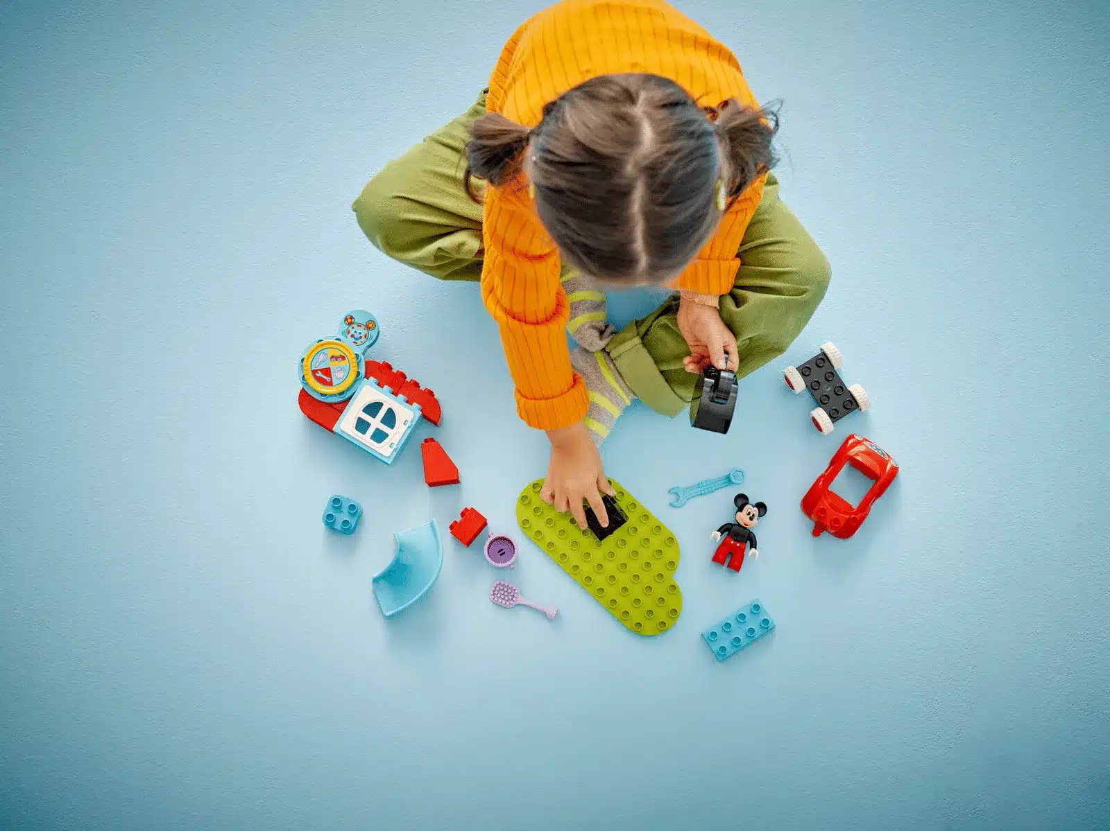 Kind baut das LEGO DUPLO Micky Maus Clubhaus mit rotem Auto und Zubehörteilen auf hellblauem Hintergrund
