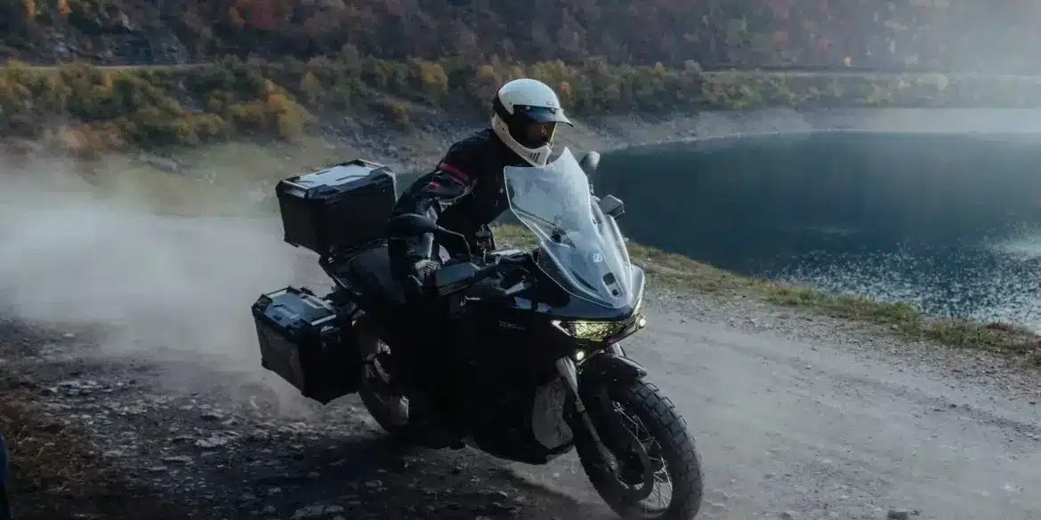 Zero DSR/X Elektro-Adventure-Bike fährt auf staubiger Schotterstraße am Seeufer entlang, herbstliche Berglandschaft im Hintergrund