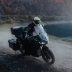 Zero DSR/X Elektro-Adventure-Bike fährt auf staubiger Schotterstraße am Seeufer entlang, herbstliche Berglandschaft im Hintergrund