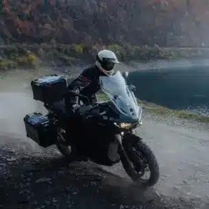 Zero DSR/X Elektro-Adventure-Bike fährt auf staubiger Schotterstraße am Seeufer entlang, herbstliche Berglandschaft im Hintergrund