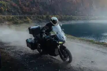 Zero DSR/X Elektro-Adventure-Bike fährt auf staubiger Schotterstraße am Seeufer entlang, herbstliche Berglandschaft im Hintergrund